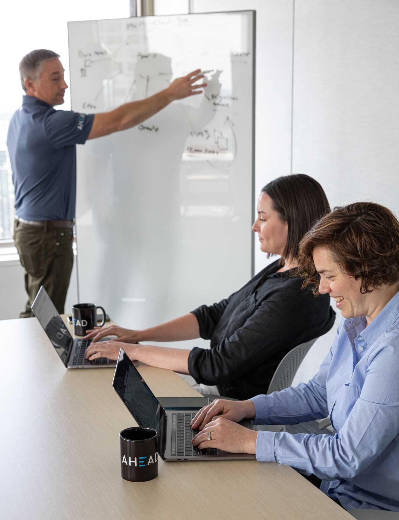 A man presenting on a whiteboard to two people on their laptops in a brightly lit office