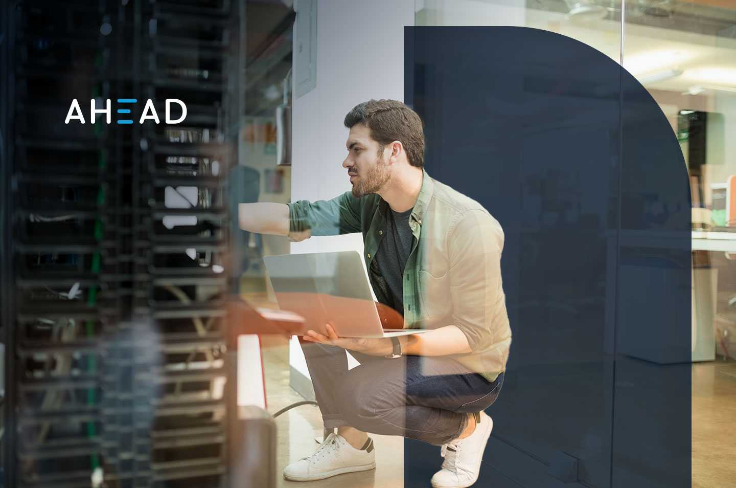 a man holding a laptop kneeling next to a data center rack